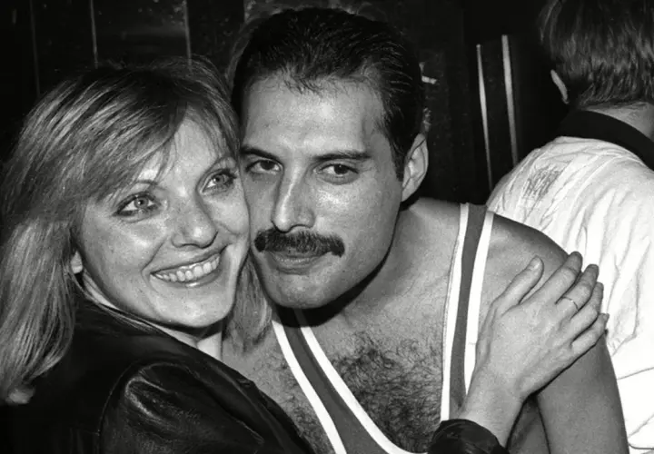 Freddie Mercury z uśmiechem, odsłaniającym jego charakterystyczne zęby, przytula się do kobiety.