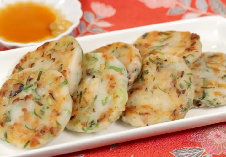 Golden-brown daikon mochi patties, studded with green onions, are served on a white platter with a small dish of dipping sauce.