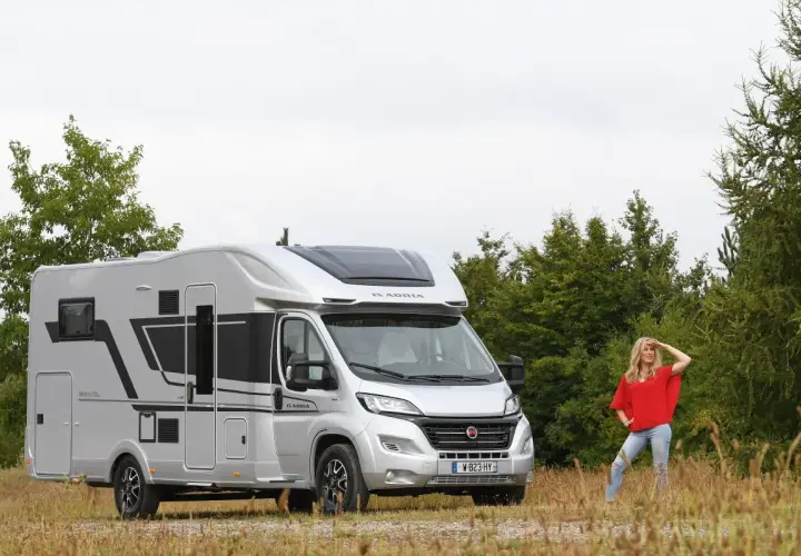 Une femme pose devant un camping-car Adria Matrix 670 SC argenté, prêt pour l'aventure.