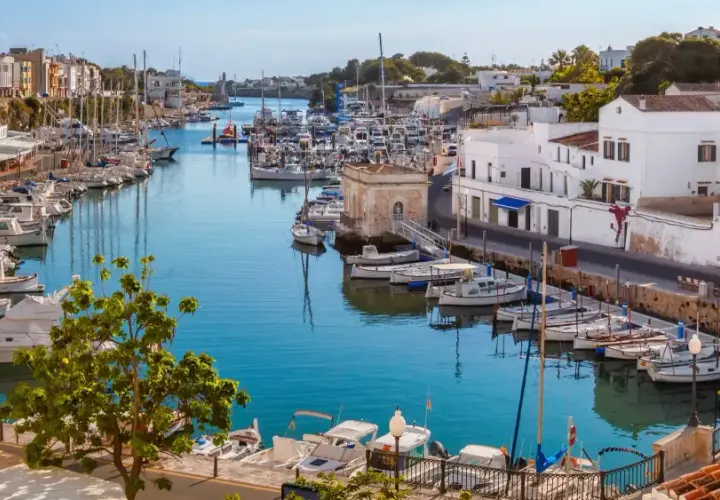 Hafen von Ciutadella, Menorca. Boote im türkisfarbenen Wasser, weiße Häuser und bunte Fassaden. Ein malerischer Blick auf die Menorca Highlights.