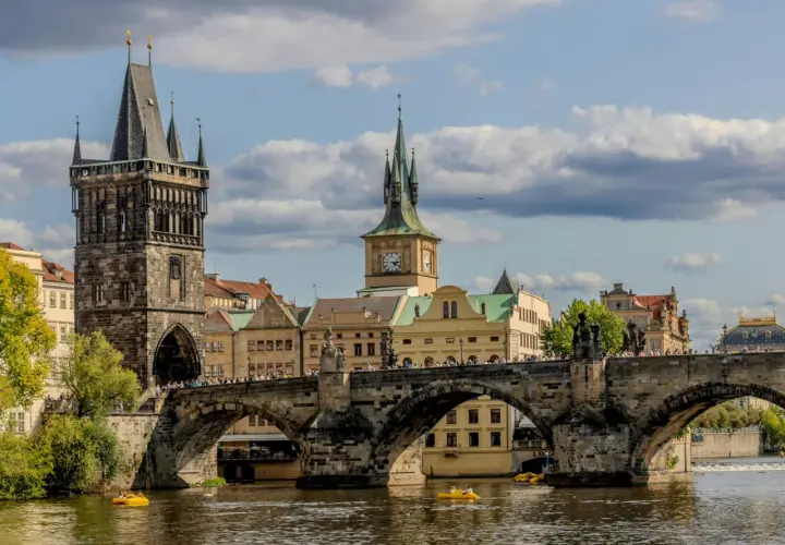 Most piękne stolice Europy, Most Karola w Pradze, z wieżami i zegarem, na tle błękitnego nieba z chmurami.