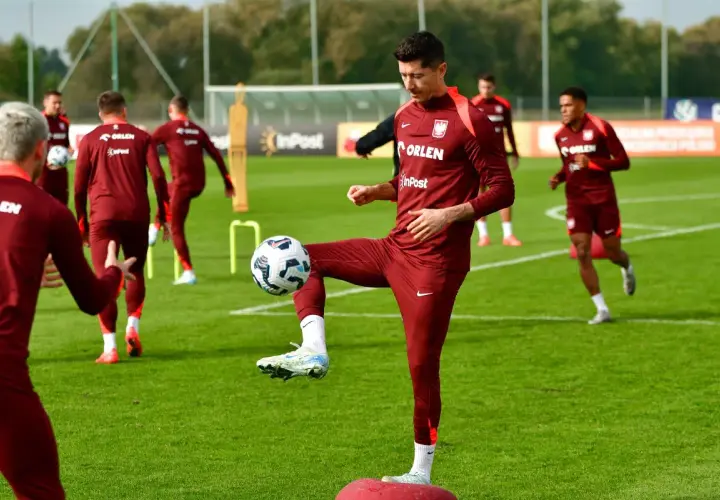 Piłkarze w bordowych strojach ćwiczą na boisku. Robert Lewandowski bawi się piłką, pokazując swoje umiejętności w najpopularniejszym sporcie na świecie.