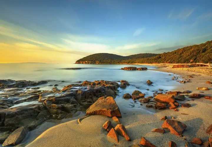 Coucher de soleil sur une plage rocheuse en Maremma, Italie. L'eau calme caresse le sable doré et les rochers ocre sous un ciel aux teintes chaudes.