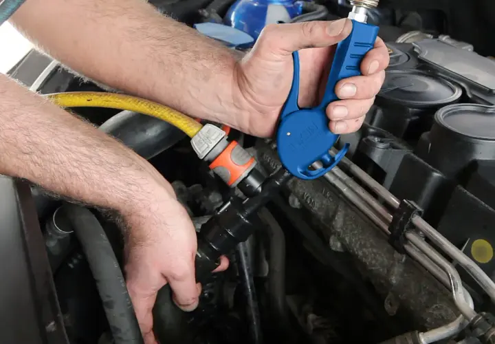 Ręce mechanika podłączają niebieskie narzędzie do węża, przygotowując środek do płukania układu chłodzenia.