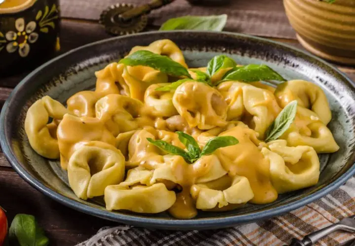 Plato de tortellini de carne cubiertos con una cremosa salsa de queso y decorados con hojas de albahaca fresca.