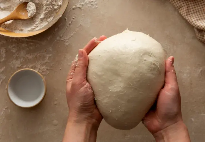 Mani che impastano un panetto soffice, ingrediente chiave per imparare come fare il lievito madre. Farina e ciotolina a lato.