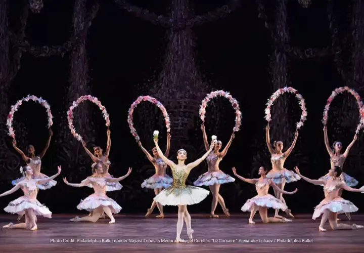 Balettdansöser i ljusa dräkter utför en scen från "Le Corsaire", med blomsterkransar i händerna.