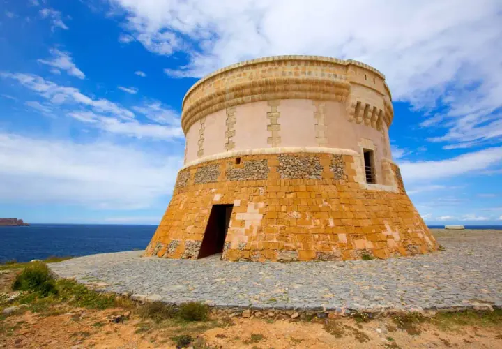 Torre de defensa costera, un lugar histórico que ver en Fornells. El mar azul y el cielo nublado completan la vista.