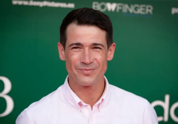 Juan José Ballesta, cuya edad se refleja en su mirada, sonríe con camisa rosa ante un fondo verde con logos.