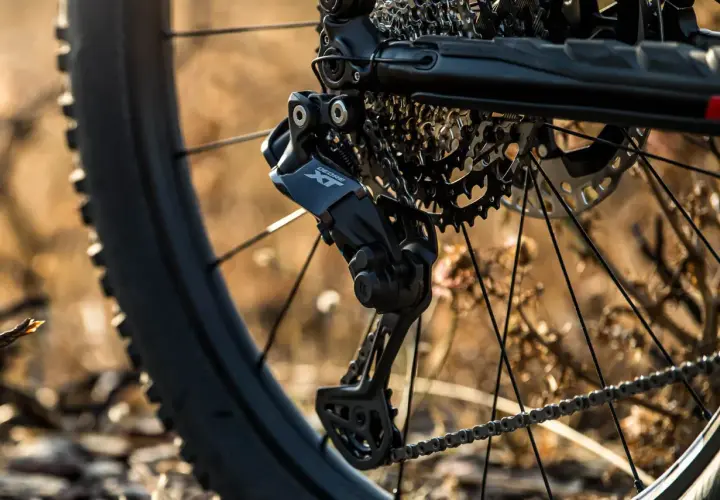 Close-up of a bicycle's rear derailleur, showcasing the Shimano Deore XT component, hinting at advanced Shimano wireless shifting technology.