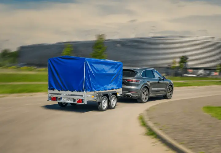 Szary SUV z przyczepą pod niebieską plandeką jedzie z dużą prędkością, mijając nowoczesny budynek.