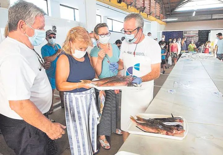 En la lonja del pescado, un hombre con delantal muestra unos peces a una mujer. Otras personas observan.