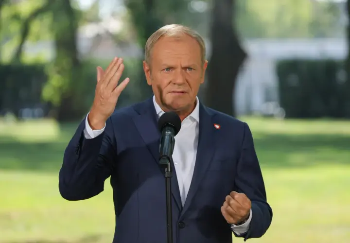 Donald Tusk w garniturze, przemawiający do mikrofonu, z serduszkiem na klapie.
