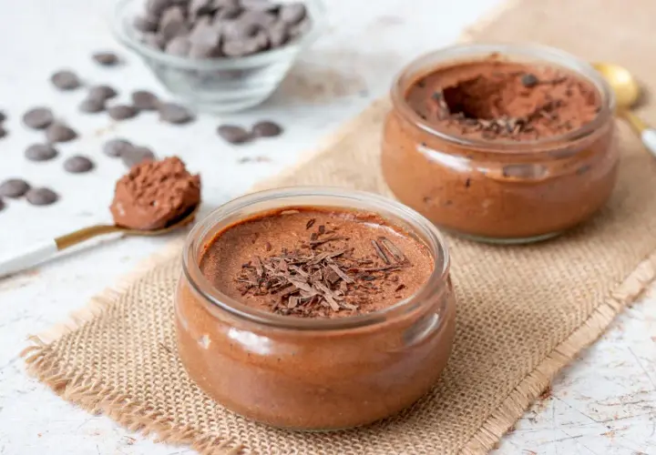 Deux pots de mousse au chocolat, l'un garni de copeaux de chocolat noir, sur une toile de jute. Une cuillère dorée présente une portion de cette délicieuse mousse.