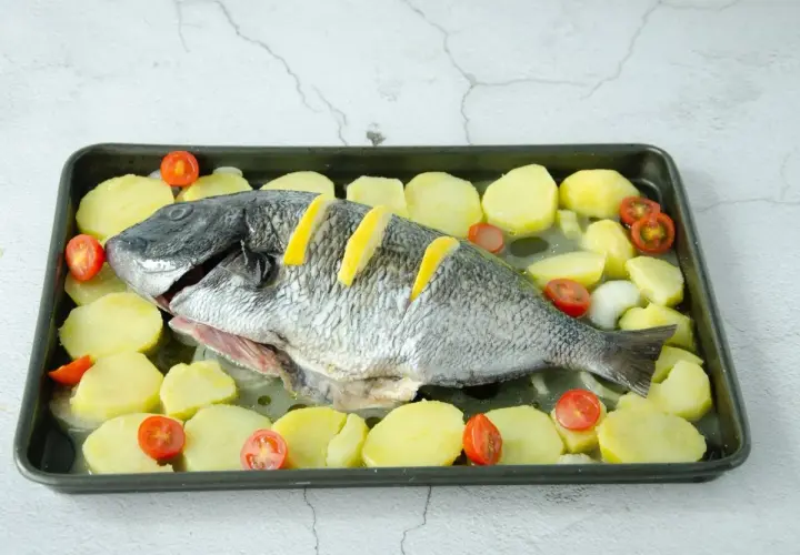 Un sargo pez entero, listo para hornear, rodeado de rodajas de patata y tomates cherry en una bandeja.
