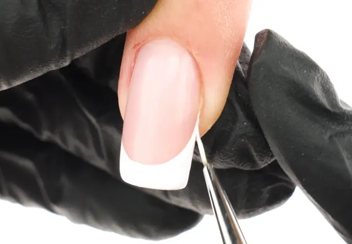Precyzyjne malowanie białej końcówki french manicure bez użycia pasków. Dłoń w czarnej rękawiczce trzyma pędzelek.