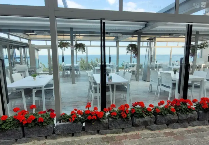 Letnia atmosfera przy plaży w Hotelu Lambert. Stoliki i krzesła czekają na gości, a morze w tle zaprasza do relaksu.