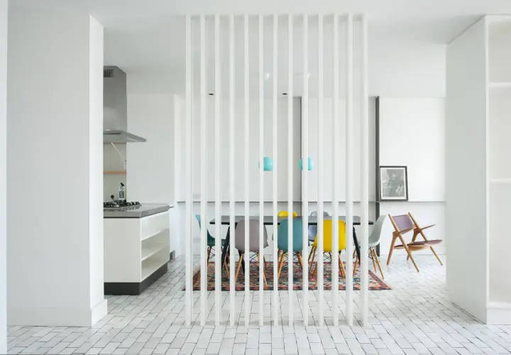 Un divisorio a listelli bianchi separa la cucina moderna da una zona pranzo con sedie colorate. Un'idea per come riempire spazio tra cucina e muro.