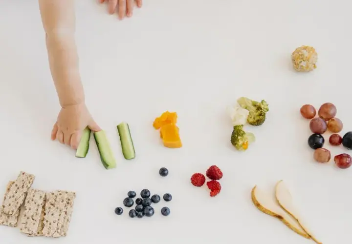 Mano di bimbo afferra bastoncini di cetriolo, parte di un pasto con tagli sicuri svezzamento: frutta, verdura e cracker.