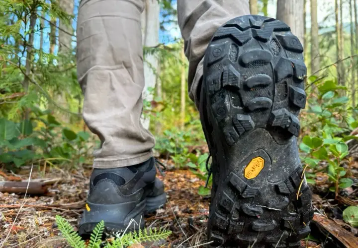 Close-up of a person's hiking boots on a forest trail, showcasing the aggressive tread pattern and the iconic yellow Vibram sole logo, highlighting the durability and grip of various Vibram sole types.