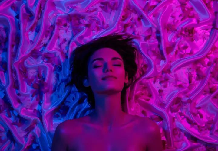 Donna con occhi chiusi, immersa in un letto di seta e petali, illuminata da luci blu e rosa. Un'immagine che evoca il piacere e il mistero del perché godiamo quando veniamo.
