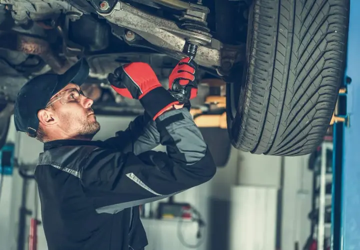 Mechanik w rękawiczkach dokręca sworzeń wahacza kluczem nasadowym pod podniesionym samochodem.