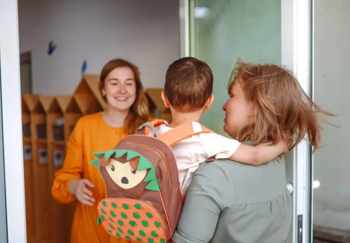 Madre abraza a su hijo con mochila de erizo, mientras una maestra sonríe. Inicio del periodo de adaptación infantil.