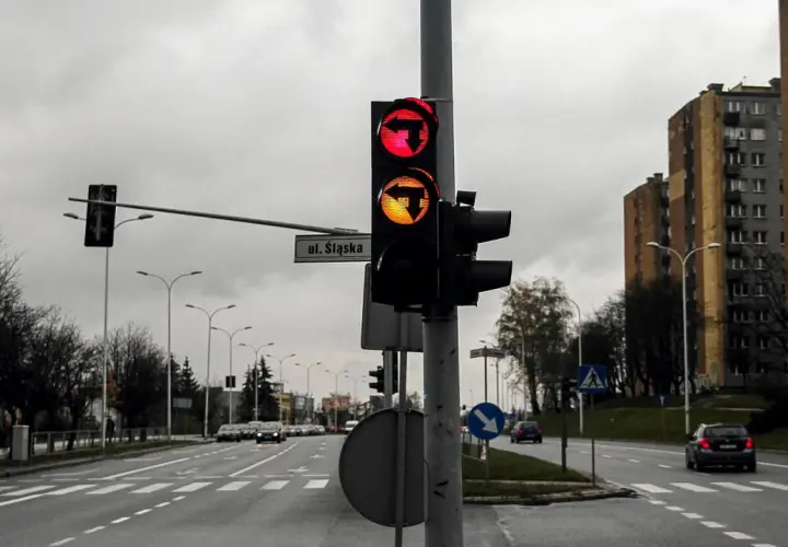 Sygnalizator S3 z czerwonym i żółtym światłem kierunkowym w lewo, na tle pochmurnego nieba i bloków mieszkalnych.