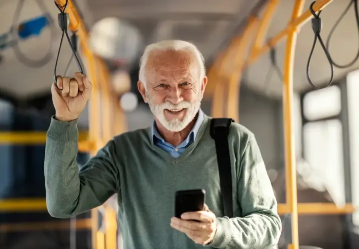 Uśmiechnięty starszy pan w autobusie, korzystający z ulgi dla emerytów w Wałbrzychu. Trzyma telefon i poręcz.
