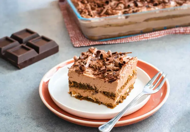 Una fetta di tiramisù cioccolato, ricca e cremosa, guarnita con riccioli di cioccolato. Accanto, pezzi di cioccolato fondente e un'intera teglia.