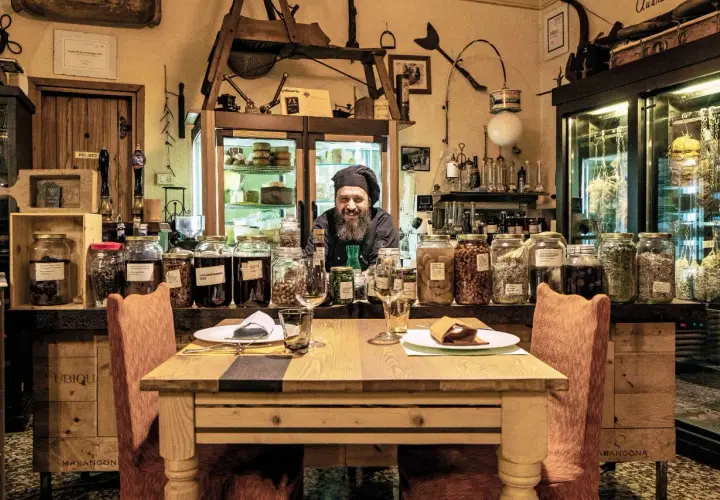 Un cuoco sorridente in un locale con salumi appesi e barattoli di conserve. Un'atmosfera unica per ristoranti particolari a Brescia e provincia.