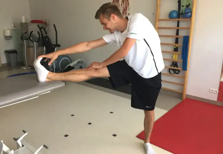 Młody mężczyzna wykonuje ćwiczenia rozciągające, próbując osiągnąć szpagat męski. W tle sprzęt rehabilitacyjny.