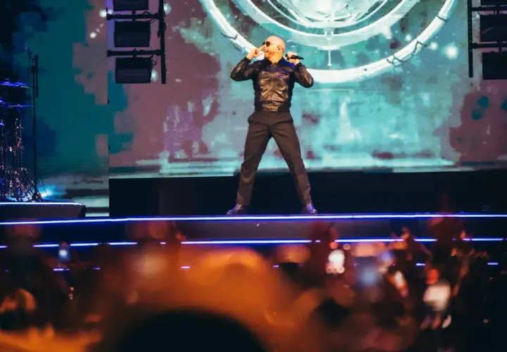 Pitbull Polska na scenie, w czarnej kurtce i spodniach, śpiewa do mikrofonu. Tłum w dole nagrywa koncert.