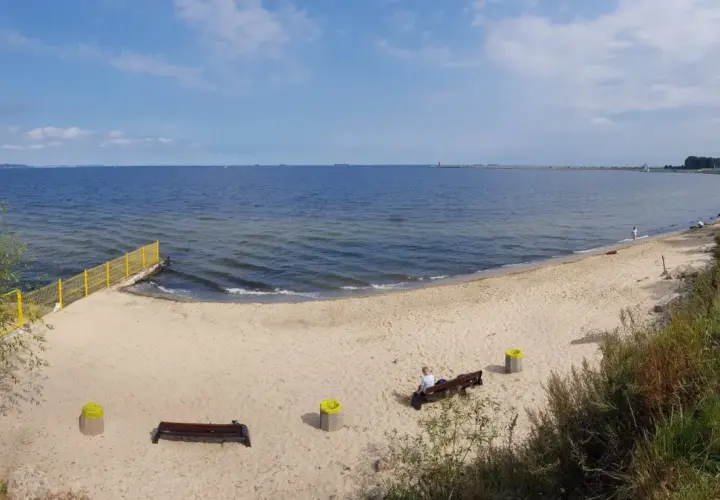 Spokojna plaża dla psów z piaskiem i morzem. Na ławce siedzi osoba, obok kosze.