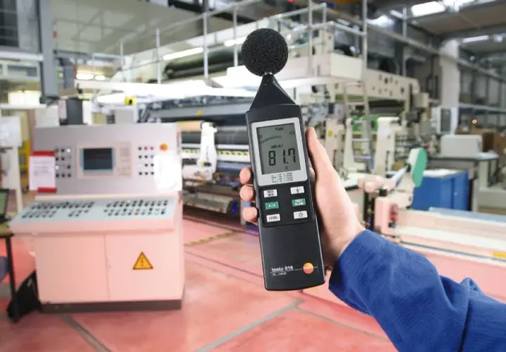 Mano sostiene sonómetro que mide 81.7 dB(A) en una fábrica, indicando el nivel de ruido.