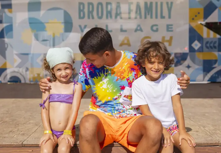 Un uomo con due bambini sorridono davanti a uno sfondo con la scritta "Brora Family Village".