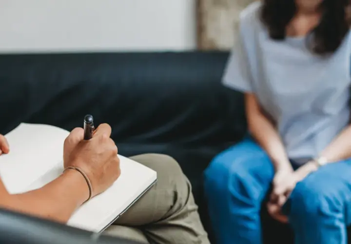 Eine Person schreibt in ein Notizbuch, während eine andere Person in blauen Jeans auf einer Couch sitzt. Dies könnte eine Szene der MDMA-Therapie sein, die bei der Heilung hilft.
