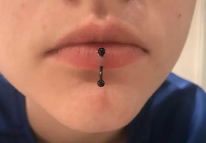 Osoba z pionowym kolczykiem w wardze (vertical labret piercing). Czarny, błyszczący kolczyk przechodzi przez środek dolnej wargi.