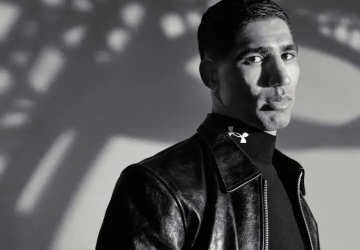 Achraf Hakimi, ein Fußballstar, blickt nachdenklich in die Kamera, trägt eine Lederjacke und ein schwarzes Rollkragenshirt mit dem Under Armour Logo.