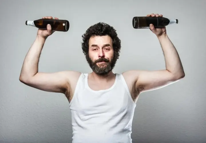 Mężczyzna w białej podkoszulce podnosi dwie butelki, symbolizując wpływ testosteronu a alkoholu na siłę i samopoczucie.