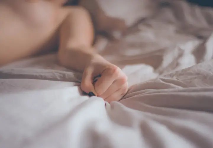 A person's hand clutches crumpled white sheets on a bed.