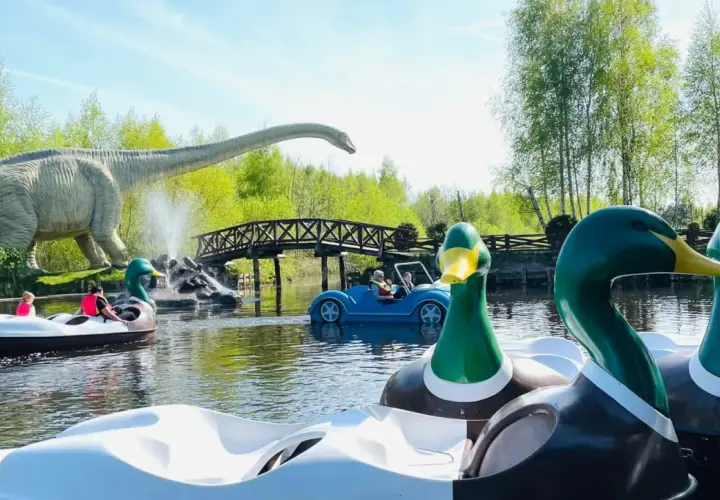 Park dinozaurów nad morzem. Dzieci pływają łódkami w kształcie kaczek i samochodu, a w tle widać gigantycznego dinozaura.