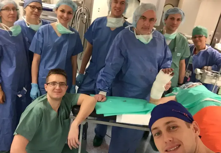 Zespół chirurgów w strojach operacyjnych pozuje do zdjęcia w sali operacyjnej, jeden z nich trzyma nogę w gipsie.