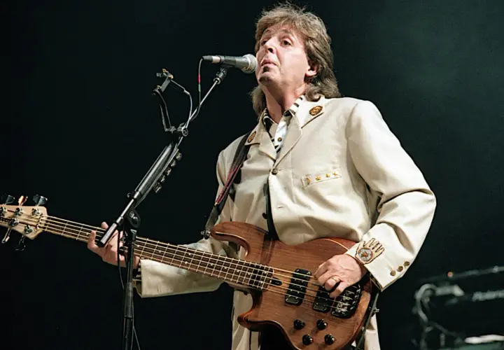 Paul McCartney z zespołu The Beatles gra na basie podczas koncertu, ubrany w elegancki beżowy garnitur.