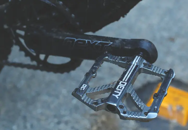 A close-up of a silver Deity pedal, ready for a Bladerunner-style descent. The crank arm reads "next SL.