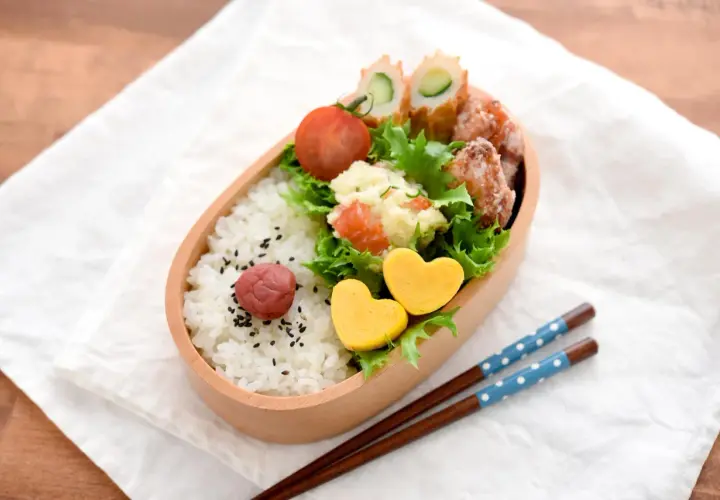 A delightful tamagoyaki bento box filled with rice, a pickled plum, potato salad, fried chicken, and cucumber rolls. Two heart-shaped tamagoyaki add a sweet touch.