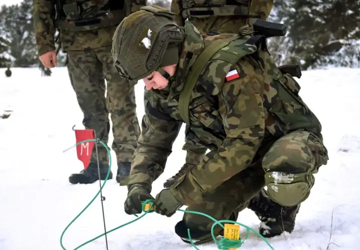 Saper w wojsku przygotowuje teren w zimowych warunkach. Żołnierz w kamuflażu pochyla się nad zielonymi przewodami, pracując ostrożnie.