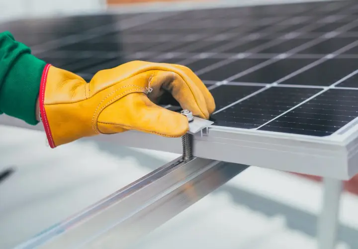 Ręka w żółtej rękawicy dokręca śrubę mocującą panel fotowoltaiczny. Instalacja PV postępuje.