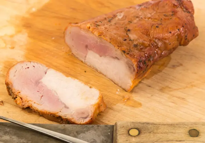 Soczysta polędwica pieczona na kanapki, pokrojona na desce, gotowa do podania. Obok nóż i widelec.