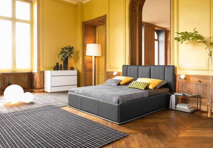 Camera da letto classica con pareti giallo ocra, letto grigio, tappeto a scacchi e arredi in legno.
